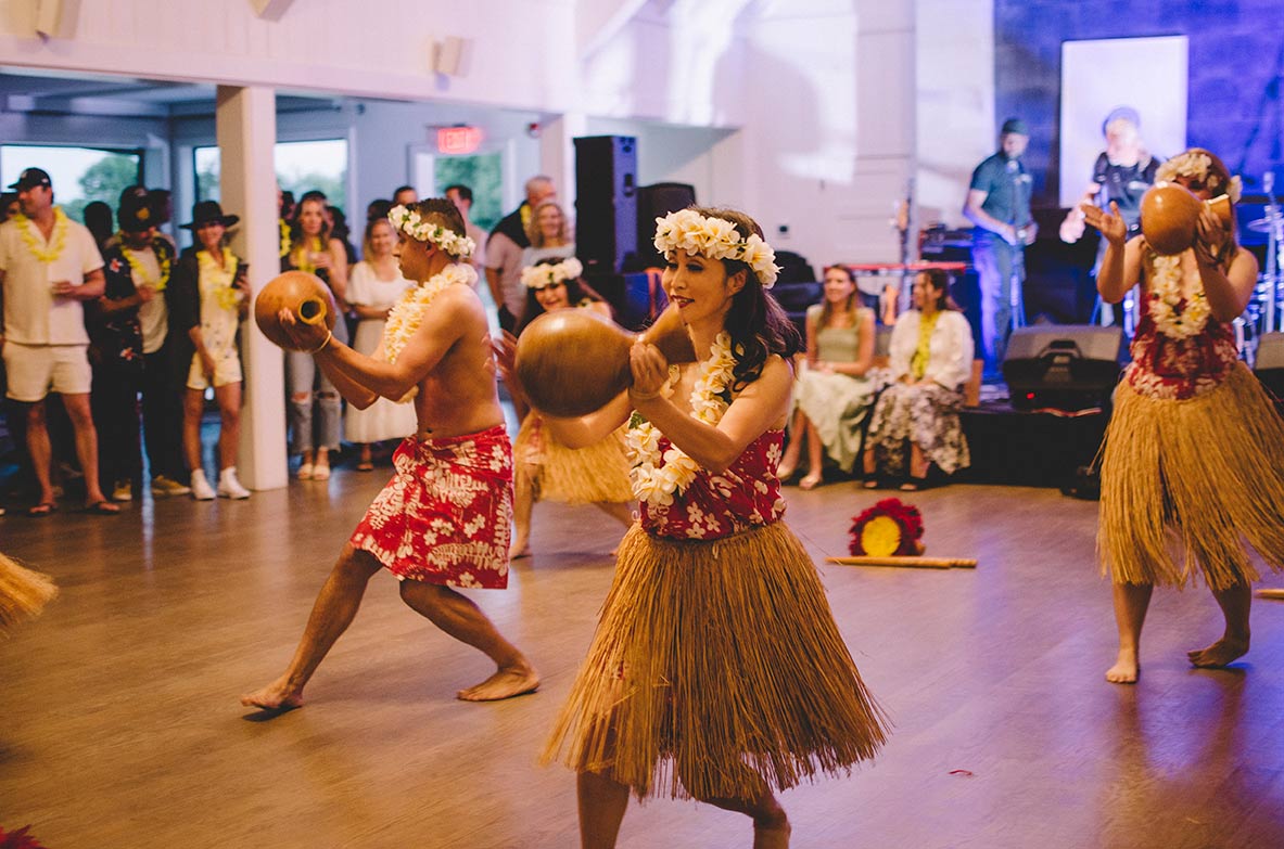 At Gurney’s, traditional hula dancers perform in an open space. In the background a standing audience looks on. 
