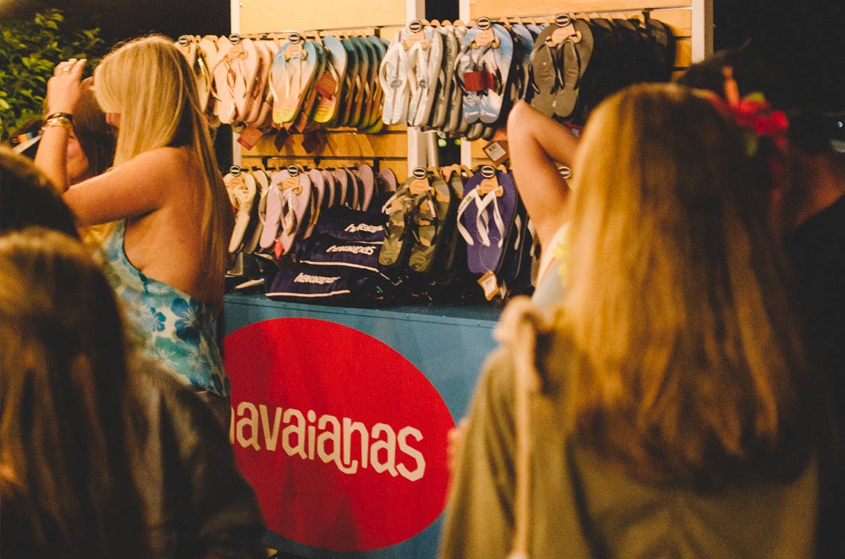 In the foreground, people stand in low light. Behind them is the Havaianas cart with rows of flip flops on display along with folded towels at Gurney's. 