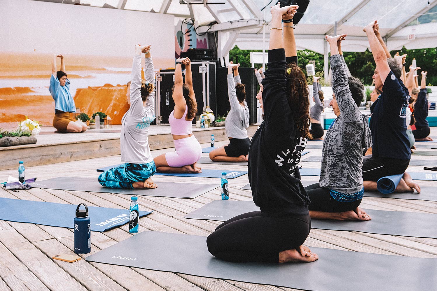 Two rows of people sit on yoga mats facing a stage and follow the lead of a yoga teacher. Everyone's hand are raised toward the air at The Surf Lodge. 