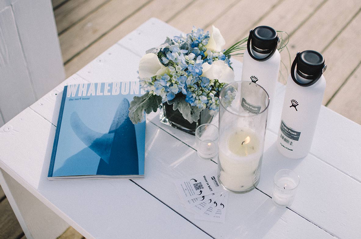 A white wood table at the surf lodge. On top is flowers, water bottles by dragon alliance and the surf issue by whalebone magazine. 