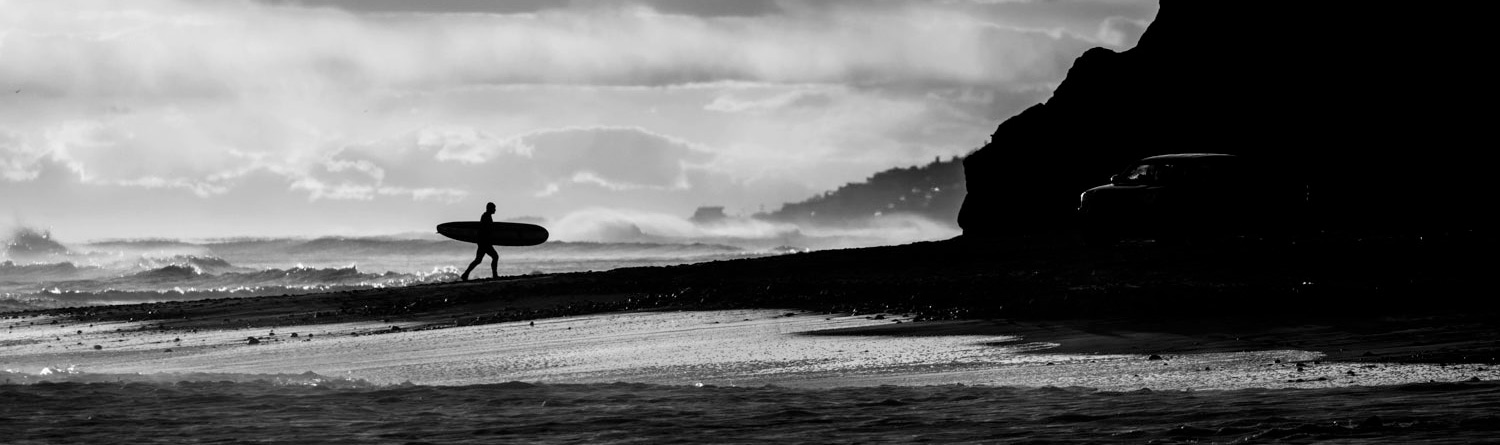 The Surf Thing. A surfer carrying a board walks toward cliffs on the right. Waves crash behind him. 