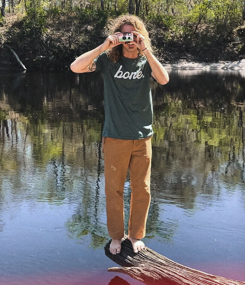 A man standing on a log above a black river takes a photo of the viewer. He's wearing a dark grey bone shirt.