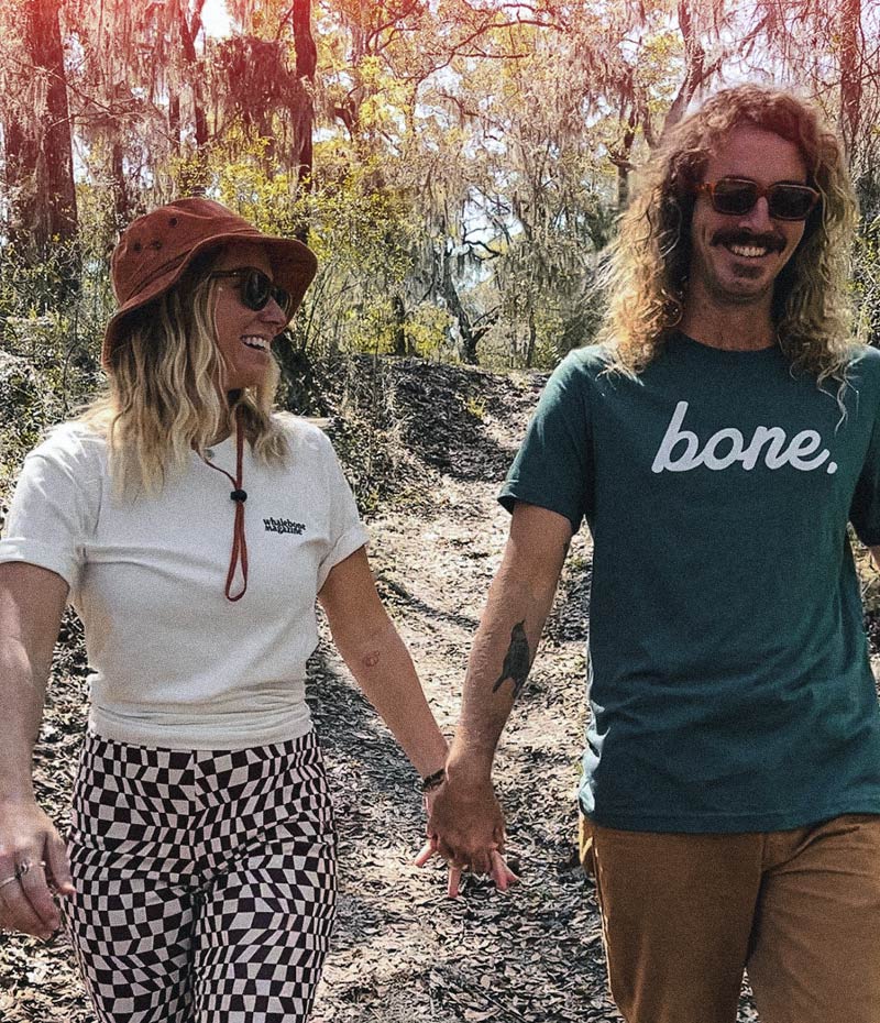 A man and a woman walk through the woods smiling. Each person is wearing a whalebone tshirt. 