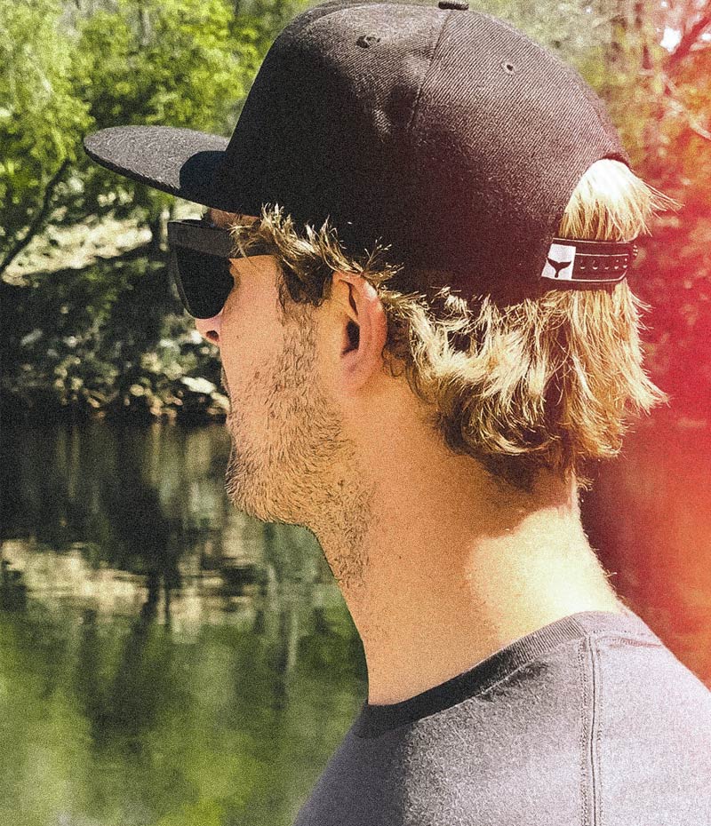 A man looking away from the viewer and toward a black river. He wears the black whalebone whaletail 5-panel hat 